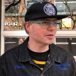 Photo of Jason Mclean in black vans baseball hat, green framed glasses and a black denim jacket